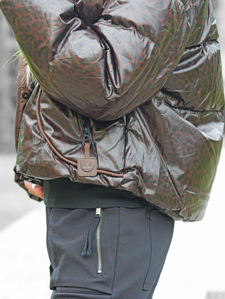 Nahaufnahme einer Person, die die OFF#DLY Winterjacke Cortina schoko trägt. Die Jacke hat ein glänzendes Leopardenmuster mit sichtbarem Reißverschluss und Lederetikett, kombiniert mit dunklen Hosen mit Reißverschlusstasche.