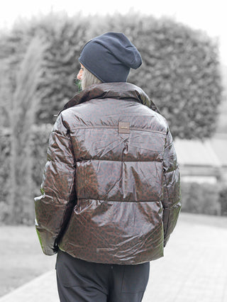 Person von hinten auf einem Parkweg, trägt die OFF#DLY Winterjacke Cortina schoko mit modernem Leoparden-Muster, kombiniert mit einer dunklen Strickmütze, umgeben von gepflegten Hecken.
