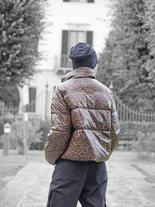 Person von hinten in einem glänzenden, leopardenmusterigen OFF#DLY Winterjacke Cortina taupe, dunkler Mütze und Cargo-Hose, stehend auf einem gepflasterten Weg vor einem weißen Gebäude mit Gittern.