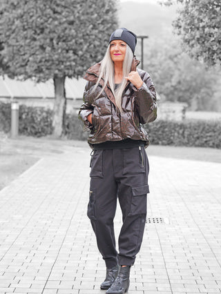 Frau trägt die OFF#DLY Winterjacke Cortina schoko, eine glänzende, gesteppte Kurzjacke mit sportlichem Schnitt, kombiniert mit schwarzen Cargo-Hosen und Stiefeln, in einem Park spazierend.