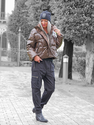 Eine Person trägt die OFF#DLY Winterjacke Cortina taupe, eine glänzende, leopardengemusterte Pufferjacke, kombiniert mit schwarzen Cargo-Hosen und einer dunklen Mütze, während sie den Kragen anpasst.