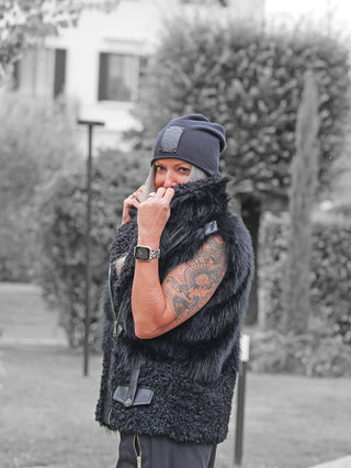 Erwachsener im Freien trägt die OFF#DLY Fell Imitat Weste Gröden black mit hohem Kragen, kombiniert mit einer dunklen Mütze und Armbanduhr, zeigt einen Drachen-Tattoo am rechten Arm.