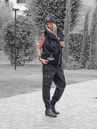 Frau im Freien trägt die OFF#DLY Fell Imitat Weste Gröden black mit asymmetrischem Reißverschluss, kombiniert mit lockeren Cargo-Joggern und Stiefeletten, betont durch ihre Tattoos und Accessoires.