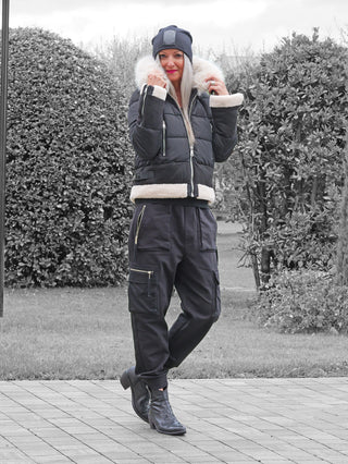 Eine Frau trägt die OFF#DLY Jacke Canazei black mit Kapuze und hellem Kunstfellbesatz. Sie steht draußen, gekleidet in eine stilvolle, quilted Winterjacke, ideal für kalte Tage.