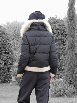 Person trägt die OFF#DLY Jacke Canazei black, eine schwarze, gesteppte Winterjacke mit hellem Kunstfellbesatz an der Kapuze, im Garten stehend, ideal für stilvolle Wärme im Winter.