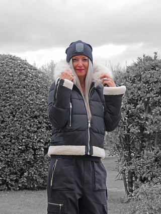 Frau im Freien mit der OFF#DLY Jacke Canazei black, einer schwarzen, gesteppten Winterjacke mit cremefarbenem Kunstfell an Kapuze, Saum und Ärmeln, hält den Fellkragen fest.