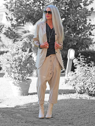 Person in einem Garten trägt die OFF#DLY Baggy Pant Doreen stone mit lockerer Passform und Knopfleiste, kombiniert mit passendem Blazer, kniehohen Stiefeln und dezentem Schmuck für einen modernen Look.