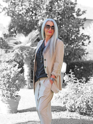 Die Frau trägt den OFF#DLY Blazer Abigail Stone in leichtem Oversize-Schnitt, kombiniert mit einer schwarzen Oberteil und passender Hose, ergänzt durch Sonnenbrille und Schmuck, im Garten stehend.