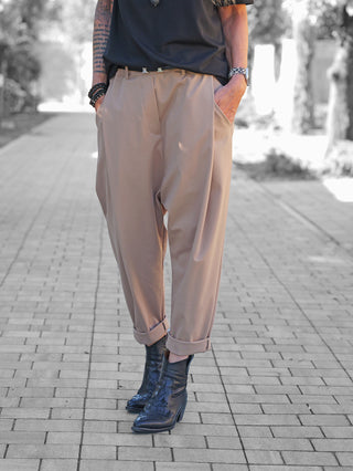 Die OFF#DLY Baggy Pant Leyla beige zeigt sich lässig-modern mit lockerem Schnitt und umgeschlagenem Saum, getragen von einer Person auf einem gepflasterten Weg, kombiniert mit schwarzen Lederstiefeln.