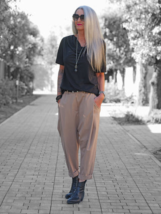 Schlanke Frau trägt OFF#DLY Baggy Pant Leyla beige, locker geschnitten mit hochgekrempeltem Saum, kombiniert mit schwarzem T-Shirt. Perfekt für einen lässigen, modernen Look auf einem Baumweg.