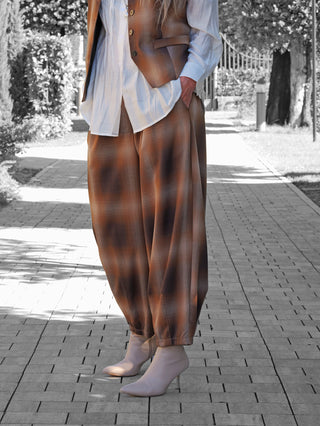 Person trägt OFF#DLY Checker Pant Dunja braun: Weit geschnittene braun-karierte Hose mit Bundfalten, kombiniert mit beiger Stiefelette, betont urban-eleganten Stil auf gepflastertem Weg.