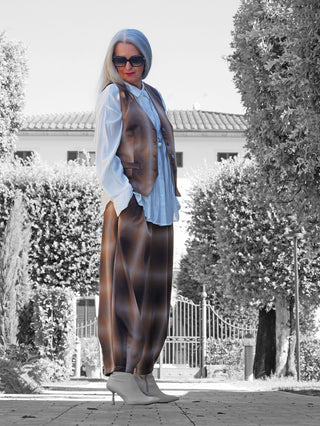 Frau trägt OFF#DLY Checker Pant Dunja braun, weite Karohose mit Bundfalten, kombiniert mit weißer Bluse und Stiefeln. Eleganter, lässiger Look mit moderner Karomuster-Optik.