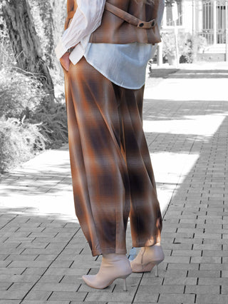 Person trägt OFF#DLY Checker Pant Dunja braun, weite karierte Hose mit Bundfalten, kombiniert mit weißem Hemd und brauner Weste, dazu beigefarbene, spitze Stiefeletten auf einem Gehweg.