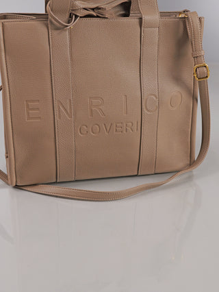 Enrico Coveri Shopper taupe aus beigefarbenem, strukturiertem Leder mit geprägtem Logo. Ausgestattet mit flachen Henkeln und verstellbarem Schulterriemen mit goldfarbener Schnalle, präsentiert auf glänzender Oberfläche.
