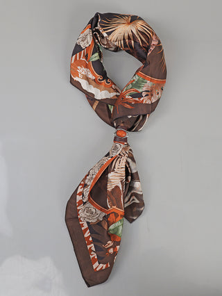 Das Bild zeigt das OFF#DLY Tuch Oak mit Spange brown, ein quadratisches Seidentuch mit tropischen Blättern und Blumen, in Brauntönen, Orange und Grün, dekorativ durch eine Spange drapiert.