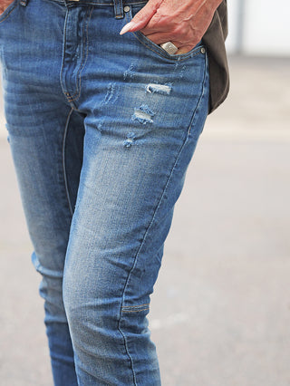 OFF#DLY Denim Bergamo midblue: Schmal geschnittene, distressed Jeans mit Rissen und abgenutzten Stellen am Oberschenkel. Hand in Tasche, Ring sichtbar, auf gepflastertem Untergrund.