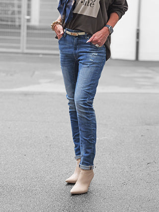 Person trägt OFF#DLY Denim Bergamo midblue Jeans mit schmalem Schnitt, leichten Used-Effekten und leicht aufgerollten Säumen, kombiniert mit cremefarbenen Stiefeletten, einem hellen Gürtel und einem locker sitzenden Oberteil.