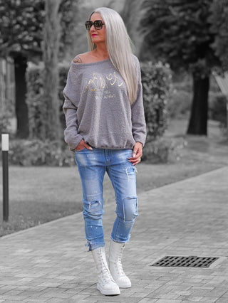 Erwachsene Frau trägt das OFF#DLY Teddy Shirt grau, ein lockeres, off-shoulder, kuscheliges Oberteil mit goldener Schrift, kombiniert mit Jeans und weißen Stiefeln im Park.