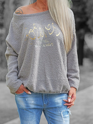 Frau trägt das OFF#DLY Teddy Shirt grau, ein kuscheliges, off-shoulder Pullover mit goldener Schrift, kombiniert mit hellblauen Jeans. Eine Hand in der Tasche, die andere trägt Ringe.