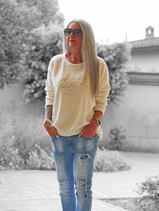 Ältere Frau trägt das OFF#DLY Teddy Shirt beige mit goldenen Buchstaben, kombiniert mit hellblauen Jeans, Sonnenbrille und Schmuck, vor einem verschwommenen Gartenhintergrund.