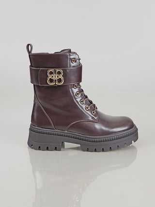 OFF#DLY Boot BB/L brown aus glattem, poliertem Leder mit Schnürung, goldener Schnalle am Schaft, dicke Gummisohle und hinterer Zuglasche, präsentiert auf einer reflektierenden Oberfläche.