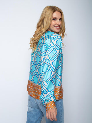 Person trägt die Emily Bluse 156196 cognac mit blau-weißem abstraktem Print und geometrischen Akzenten in Orange. Dazu hellblaue Jeans. Hemd mit kleinen Stehkragen und Manschetten an den Ärmeln.