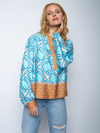 Emily Bluse 156196 cognac: Frau trägt locker sitzende Bluse mit geometrischem Muster, aqua-blauem Hauptfeld, orange-weißen Scroll-Details, kombiniert mit hellen Jeans. Fließender Viskose-Satin, kleiner Stehkragen, lange Ärmel.