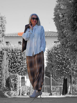 Frau in einer stilvollen, oversized OFF#DLY Bluse Pordoi white mit Falten, kombiniert mit braunen, weiten Hosen und grauen Stiefeletten, vor einem desaturierten Gartentor und Villa lächelnd.