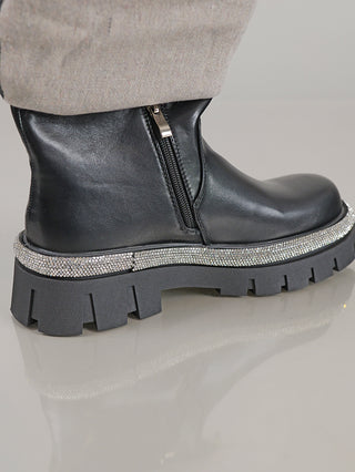 Nahaufnahme des OFF#DLY Boot Star black, einem schwarzen Lederstiefel mit klobiger Gummisohle, seitlichem Reißverschluss und glitzernden Strassverzierungen entlang der Sohle. Der Saum einer hellen Hose ist sichtbar.