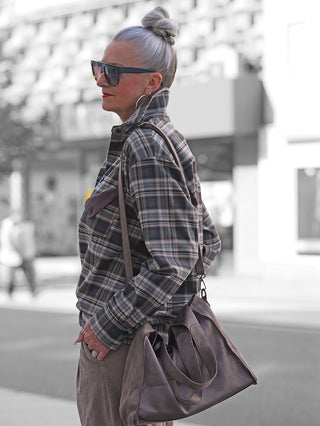 Eine Person trägt die Lumina Bluse braun Karo im Oversize-Stil, kombiniert mit silbrigen Haaren in einem Dutt, großen Sonnenbrillen und einem taupefarbenen Ledertasche auf einer städtischen Straße.