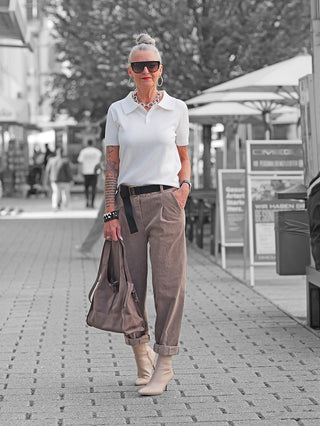 Ältere Frau trägt OFF#DLY Cord Pant Lavinia taupe mit hohem Bund und schwarzem Gürtel, kombiniert mit weißem Polo und cremefarbenen Stiefeletten, während sie eine taupefarbene Ledertasche hält.