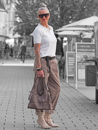 Ältere Frau trägt OFF#DLY Cord Pant Lavinia taupe, hoch tailliert, mit weitem Bein und Cordstruktur. Kombiniert mit weißem Polo und Accessoires, hält sie eine braune Ledertasche.