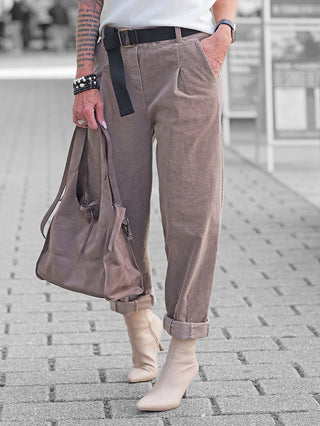 Person trägt die OFF#DLY Cord Pant Lavinia taupe mit umgeschlagenen Bündchen, kombiniert mit einem schwarzen Webgürtel, cremefarbenen Stiefeletten und einer braunen Leder-Tote in der Hand.