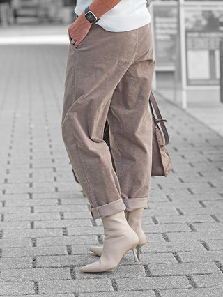 Person trägt OFF#DLY Cord Pant Lavinia taupe, locker geschnittene Cordhose mit umgeschlagenen Bündchen, kombiniert mit beigen Stiefeletten. Linke Hand in Tasche, Smartwatch am Handgelenk.