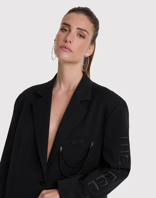 Eine junge Frau trägt den ALIX Grandad Blazer black, einen oversized schwarzen Blazer mit Stickerei ALIX und abnehmbarer Perlenkette an der Brusttasche, sowie THE REBEL am Ärmel.