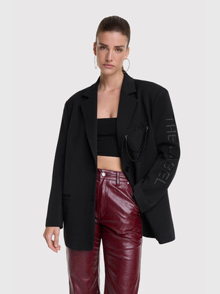 Modell trägt den ALIX Grandad Blazer black mit Oversized-Fit, Schulterpolstern, abnehmbarer Perlenkette an der Brusttasche und „THE LABEL“-Schriftzug am Ärmel. Kombiniert mit burgunderroten Lederhosen.