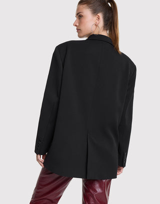 Frau in ALIX Grandad Blazer schwarz, oversized mit Schulterpolstern und Perlenkette an Brusttasche, trägt burgundy Leder-Look-Hosen. Blazer-Stickereien und große Ohrringe sichtbar. Haare im Pferdeschwanz.