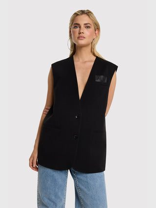 ALIX Oversized Weste black getragen von einer jungen Frau, ärmellos mit tiefem V-Ausschnitt und Brusttasche, kombiniert mit hellen Jeans mit Nieten und schlichten Ohrringen, minimalistischer Look.