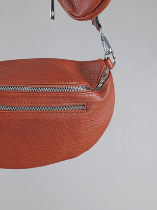 Nahaufnahme der OFF#DLY Crossbody Bag zimt, einer kleinen, halbmondförmigen Gürteltasche aus genarbtem Leder mit zwei silbernen Reißverschlüssen, Drehverschluss und D-Ring am verstellbaren Riemen.