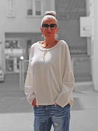 Frau trägt das OFF#DLY Sweat Elisa white, ein locker sitzendes, langärmeliges weißes Oberteil mit raffiniertem Rückenausschnitt, kombiniert mit urbanem Schmuck und Jeans auf einer Stadtstraße.