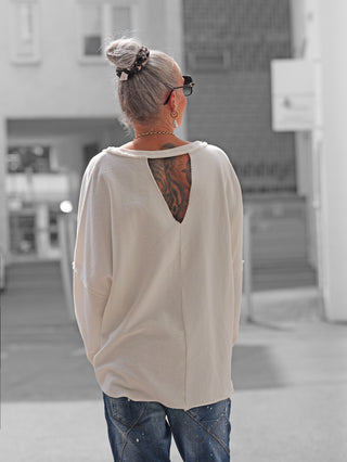 Person von hinten in urbaner Umgebung, trägt das OFF#DLY Sweat Elisa white mit Dreiecks-Rückenausschnitt, grauem Dutt mit Scrunchie, Sonnenbrille, goldene Ohrringe, Kette und locker sitzende, farbgesprenkelte Jeans.