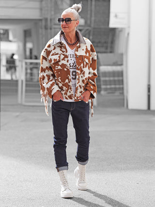 Eine ältere Person trägt die auffällige Lumina Jacke brown/white mit Kuhfleckenmuster und Fransen, kombiniert mit einer weißen Grafik-T-Shirt, dunklen Jeans und weißen Stiefeln auf einem städtischen Gehweg.