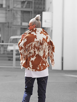 Eine Person trägt die Lumina Jacke brown/white mit Kuhfleckenmuster und langen Fransen. Die Jacke verleiht ein auffälliges Western-Flair, kombiniert mit einer weißen T-Shirt-Schicht und dunklen Hosen.