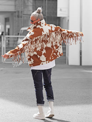 Person von hinten in der Stadt, trägt die Lumina Jacke brown/white mit Kuhmuster und Fransen, kombiniert mit hochgekrempelten Jeans und weißen Stiefeln, Haare im Dutt, Sonnenbrille.