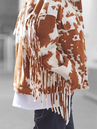 Die Lumina Jacke brown/white präsentiert sich mit Kuhfleckenmuster, langen Fransen an Ärmeln und Saum, sichtbaren Klappentaschen, über einem weißen Shirt und kombiniert mit dunklen Jeans im Western-Stil.