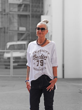 Frau in OFF#DLY Longsleeve Boston white Sweatshirt mit sportlichem Boston-Print, lässigem Schnitt kombiniert mit Jeans und Accessoires, verkörpert entspannten College-Stil in urbaner Umgebung.