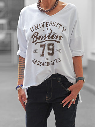 Person trägt das OFF#DLY Longsleeve Boston white mit sportlichem Boston-Print, kombiniert mit dunkelblauer Jeans. Accessoires: klobige Halskette, Smartwatch, blaue Armband und Ringe. Lockerer College-Stil für lässige Outfits.
