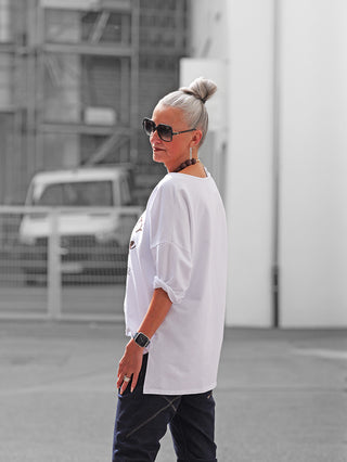 Frau trägt OFF#DLY Longsleeve Boston white, locker sitzendes weißes Shirt mit sportlichem Boston-Print, lässig kombiniert mit dunklen Hosen, Accessoires und Sonnenbrille in urbaner Umgebung.