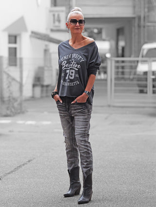 Frau trägt das OFF#DLY Longsleeve Boston anthrazit mit University Boston 79 Massachusetts-Print, lässig über die Schulter, kombiniert mit schmalen Jeans und Stiefeletten, urbaner Hintergrund unscharf.