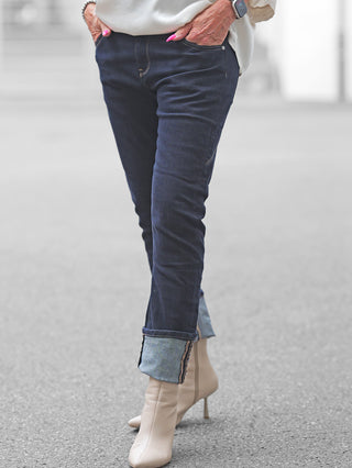 Tragende Person mit OFF#DLY Dark Denim Luino, umgeschlagene Säume zeigen hellere Innenseite. Kombiniert mit cremefarbenen Stiefeletten, silbernem Armband und Smartwatch. Perfekte Jeans für elegante oder lässige Looks.